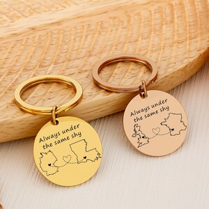 Puede incluir: Dos llaveros con colgantes redondos. Uno es dorado, el otro es oro rosa. Cada colgante tiene un contorno de mapa con un corazón y el texto "Always under the same sky".