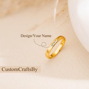 May include: A gold-colored ring with the name "Grace" engraved on it, along with an infinity symbol. The ring is displayed on a soft, neutral background. The text "Design/Your Name" and "CustomCraftsBy" are also visible.