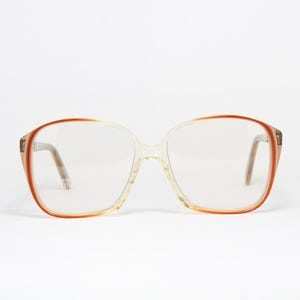 Könnte beinhalten: Vintage Brille mit einem transparenten, übergroßen Rahmen. Die Rahmen haben einen Farbverlauf, der von einem hellen Beige zu einem rötlich-braunen Farbton übergeht. Die Bügel sind hellbraun. Die Brille ist vor einem weißen Hintergrund platziert.