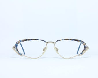 May include: Eyeglasses with a gold-colored metal frame and blue lenses. The temples feature a patterned design in shades of blue, brown, and white. The glasses are set against a white background.