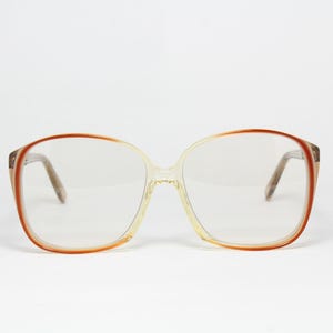 Könnte beinhalten: Eine Brille mit großen, abgerundeten Rahmen, die ein durchscheinendes beiges Inneres und einen kräftigen, rötlich-orangen Außenrand aufweisen. Die klaren Gläser sind in die breite, leichte Kunststoffstruktur mit integrierten Nasenpads eingesetzt.