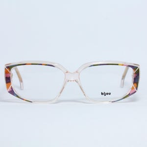 Könnte beinhalten: Brille mit transparentem Rahmen und einem geometrischen Muster in Rosa, Orange und Lila an den äußeren Rändern. Der Markenname "bijou" ist auf dem unteren rechten Glas aufgedruckt. Die Brille hat eine rechteckige Form.