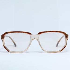 Könnte beinhalten: Klare Brille mit hellbraunem Rahmen und Bügeln. Die Fassungen haben eine rechteckige Form mit abgerundeten Kanten. Die Gläser sind klar, und die Brille ist vor einem weißen Hintergrund platziert.