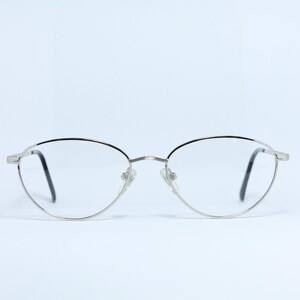 Könnte beinhalten: Brille mit silbernem Metallrahmen in ovaler Form. Die Gläser sind klar, die Bügel schwarz. Der Rahmen ist aus dünnem, glänzendem Silbermetall. Das Design ist schlicht und elegant, für den täglichen Gebrauch geeignet. Die Rahmenbreite beträgt ca. 13 cm.