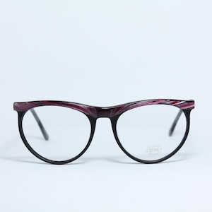 May include: A pair of eyeglasses with a black frame and clear lenses. The upper frame has a burgundy and pink marbled pattern. The glasses have a cat-eye shape and are set against a white background.