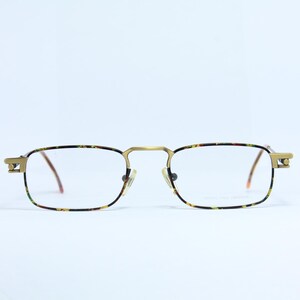 May include: Rectangular spectacles with a gold-coloured frame and patterned rims. The lenses are clear, and the arms are thin and brown. The glasses have a classic, vintage design.