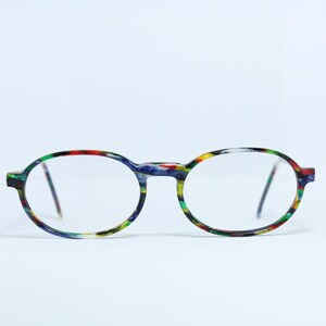 Puede incluir: Gafas de forma ovalada con un patrón moteado colorido. Las monturas presentan una mezcla de tonos azules, verdes, rojos y amarillos. Las lentes son transparentes y las varillas son finas y oscuras. Las gafas están sobre un fondo blanco liso.