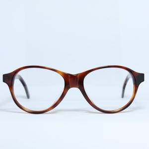 Könnte beinhalten: Eine Brille mit Schildpattmuster. Die Fassung ist warmbraun und hat eine runde Form. Die Gläser sind klar, und die Bügel sind schwarz. Die Brille ist vor einem weißen Hintergrund platziert.