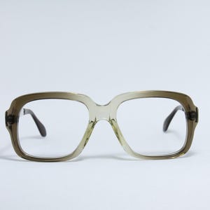 Peut inclure: Une paire de lunettes vintage avec une monture vert olive clair et des branches foncées. Les montures carrées ont un pont en trou de serrure et des verres transparents. Les lunettes sont sur fond blanc.