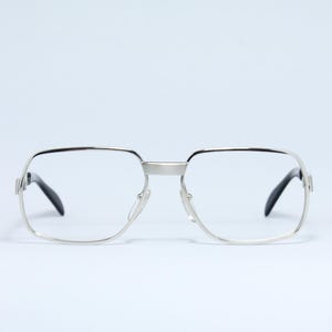 Könnte beinhalten: Brille mit silbernem Rahmen, quadratischer Form und klaren Gläsern. Die Brille hat schwarze Bügel und Nasenpads. Das Design ist ein klassischer Vintage-Stil, geeignet für den täglichen Gebrauch.
