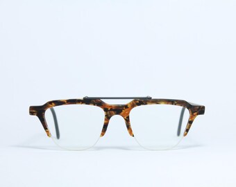 Puede incluir: Gafas con montura con estampado de carey y puente negro. Las lentes son transparentes y tienen un diseño de media montura. Las gafas tienen un estilo retro y vintage.