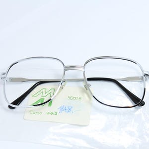 May include: Silver-rimmed glasses with clear lenses and black arms. The glasses are on a white surface with a green and white label that says "M Corso weiß 56018 14.8."