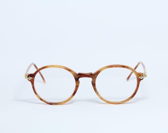 Puede incluir: Gafas redondas con un patrón de carey. Las monturas son una mezcla de tonos marrones y beige, con lentes transparentes. Las patillas son finas y se extienden hasta los terminales. Las gafas están sobre un fondo blanco.
