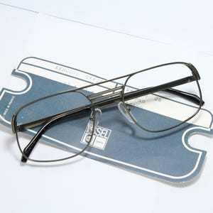 Könnte beinhalten: Silberfarbene Metallbrille mit rechteckigem Gestell und klaren Gläsern. Die Brille liegt auf einer blauen und weißen Papierkarte mit dem aufgedruckten Text "MADE IN FRANCE" und "DIESEL".