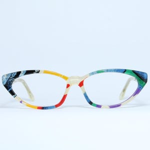 Könnte beinhalten: Eine Brille mit einem farbenfrohen, gemusterten Rahmen. Die Cat-Eye-Fassung zeigt eine Mischung aus Blau, Grün, Gelb, Rot und Lila. Die Gläser sind klar, und die Bügel sind hellbeige. Ein modisches Accessoire.