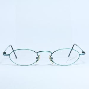 Könnte beinhalten: Oval geformte Brille mit einem türkis farbenen Metallrahmen und dünnen schwarzen Bügeln. Die Gläser sind klar, und der Rahmen hat einen kleinen Nasensteg und eine Signatur auf dem rechten Glas. Vor weißem Hintergrund.