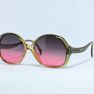 Op de afbeelding: Oversized zonnebril met een verlooplens die overgaat van roze naar donkergrijs. De monturen zijn doorschijnend olijfgroen. De zonnebril heeft een retro, vintage stijl.