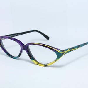 Könnte beinhalten: Cat-Eye-Brille mit lebendigem Design. Die Fassungen weisen eine Mischung aus Farben auf, darunter Lila, Türkis und Gelb, mit schwarzen Bügeln. Die Brille hat einen einzigartigen, künstlerischen Look.