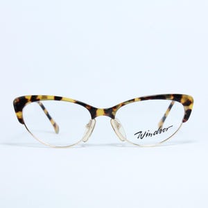 Könnte beinhalten: Cat-Eye-Brille mit Schildpattmuster auf dem oberen Rahmen und den Bügeln. Der untere Rahmen ist aus goldfarbenem Metall. Der Markenname "Windsor" ist auf dem rechten Glas aufgedruckt.