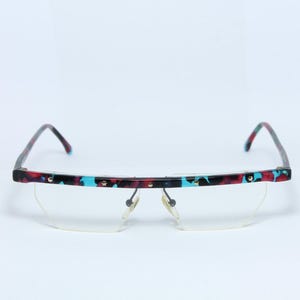 May include: Spectacles with a colourful speckled pattern on the frame. The frame features a combination of red, black, and turquoise colours. The lenses are a clear, half-rim design.