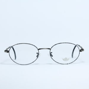 May include: Oval-shaped spectacles with a dark grey frame and clear lenses. The frame has a textured pattern. The temples are thin and curve around the ears. The glasses have a small logo on the right lens.