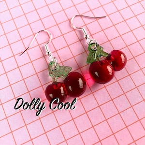 May include: A pair of silver dangle earrings with red and green cherry charms. The cherries are made of glass and have a glossy finish. The earrings are on a pink and white checkered background.