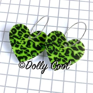 May include: A pair of heart-shaped earrings with a neon green leopard print design. The earrings are suspended from silver-colored hoops. The text "Dolly Cool" is visible in the image.