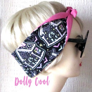 May include: A pink and black headband with a spooky gingerbread house print. The headband is tied in a knot at the top and has a black and white pattern of gingerbread houses, bats, and stars. The text "Dolly Cool" is printed in pink at the bottom of the image.