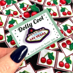 May include: A purple and white enamel pin with the text "Welcome to Fabulous Las Vegas Nevada" in a retro style font. The pin is on a white card with the text "Dolly Cool" and "Cool Jewels for Rockin' Gals".