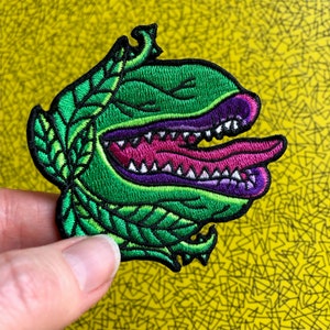 May include: Green and purple embroidered patch of a cartoon monster with a large mouth and tongue. The monster is surrounded by green leaves.
