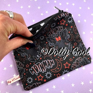 May include: Black zippered pouch with a repeating pattern of a Mothman graphic, red stars, and crescent moons. The word "Mothman" is printed in a stylized font. The pouch is being opened to reveal a polka dot interior.