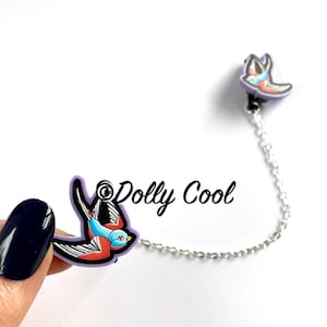 May include: A silver chain with two colourful swallow bird charms. The charms are purple, blue, red and black. The text "Dolly Cool" is visible in the image.