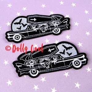 Pu&ograve; includere: Due toppe ricamate in bianco e nero raffiguranti un carro funebre con una bara, pipistrelli e una luna piena. Le toppe hanno un tema gotico e sono ambientate su uno sfondo viola con stelle bianche. Il testo "Dolly Cool" &egrave; in rosso.