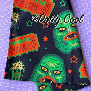 May include: Black fabric featuring a repeating pattern of green monster faces, orange stars, and movie ticket stubs that read "HORROR". Also includes popcorn graphics. The text "Dolly Cool" is visible.