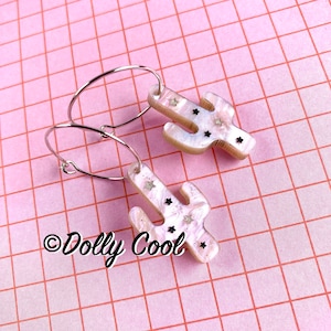 May include: A pair of silver hoop earrings with a white cactus charm. The cactus charm has a glittery finish and black stars.