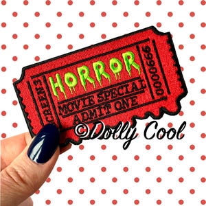 May include: A red embroidered patch resembling a cinema ticket. The patch features the word "HORROR" in dripping green letters, along with "MOVIE SPECIAL ADMIT ONE" and "SCREEN 3". The patch is held by a hand with a dark blue nail.