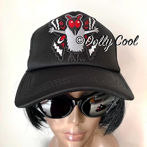 May include: A black baseball cap featuring an embroidered moth graphic with large red eyes, a grey body, and black and white wings. The text Dolly Cool is embroidered in white script on the right side of the cap above the brim.