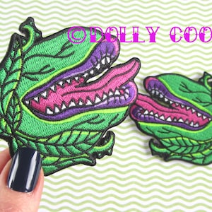 May include: Two green, purple and pink embroidered patches of a carnivorous plant with a toothy mouth and a long tongue. The patches are on a green and white striped background.