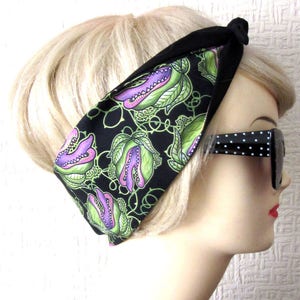 May include: A black headband with a green and purple print of carnivorous plants. The headband is tied in a knot at the top and is worn by a mannequin with blonde hair.