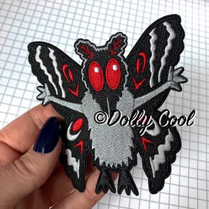 May include: An embroidered patch featuring a moth character with large red eyes, a fuzzy grey chest, and black wings accented with white wavy lines and red markings. The text Dolly Cool is embroidered in black script across the center of the moth.