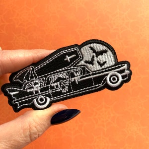May include: Black and white embroidered patch depicting a hearse with a coffin, set against a full moon and bats. This Halloween-themed patch is designed for application on clothing or accessories, with a spooky aesthetic.