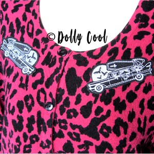 Pu&ograve; includere: Cardigan rosa con stampa leopardata, bottoni neri e due toppe ricamate. Le toppe raffigurano un carro funebre con una bara, un teschio e pipistrelli. Il testo "Dolly Cool" &egrave; visibile.