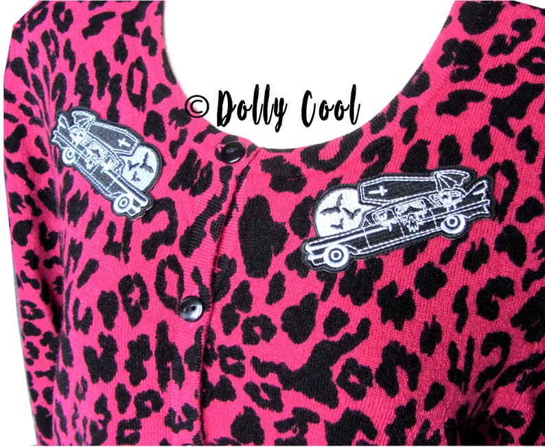 Pu&ograve; includere: Cardigan rosa con stampa leopardata, bottoni neri e due toppe ricamate. Le toppe raffigurano un carro funebre con una bara, un teschio e pipistrelli. Il testo "Dolly Cool" &egrave; visibile.
