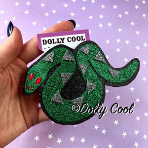 May include: A glittery green snake-shaped patch with a black border, silver triangular accents, and two red gem eyes. The patch features a purple tag with the text DOLLY COOL and the text Dolly Cool is printed in black on the lower right side.