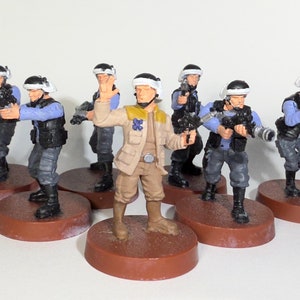 May include: A group of seven miniature figures, painted in a military style, stand on round bases. The figures are wearing blue and grey uniforms with white helmets. One figure is wearing a tan jacket and has a blue patch on their chest. The figures are holding weapons.