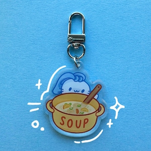 May include: A clear acrylic keychain featuring a cartoon bunny peeking over a yellow soup pot. The pot has the word "SOUP" in red and contains soup with vegetables. A silver clasp is attached to the top.