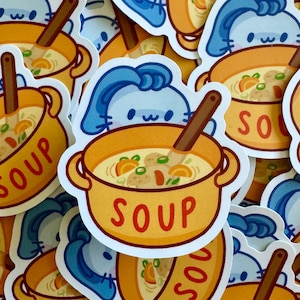 May include: A collection of stickers featuring a cartoon bowl of soup with a wooden spoon. The soup is yellow with orange and green vegetables. The word "SOUP" is written in orange on the bowl. A blue bunny character is also featured.