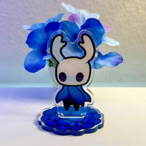 Op de afbeelding: Een heldere acrylfiguur van een stripfiguur met een witte kop, grote gebogen hoorns en een blauwe cape. Het beeldje staat op een blauwe basis met een wolkvorm. Blauwe bloemen staan op de achtergrond.