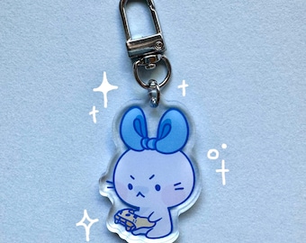 Puede incluir: Un llavero de conejito de dibujos animados azul y blanco con un lazo azul. El conejito sostiene un pequeño objeto marrón en sus patas.