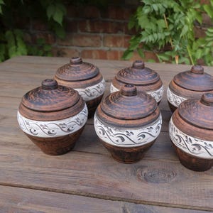 May include: Six brown ceramic jars with white floral designs and lids. The jars are arranged in a circle on a wooden surface.